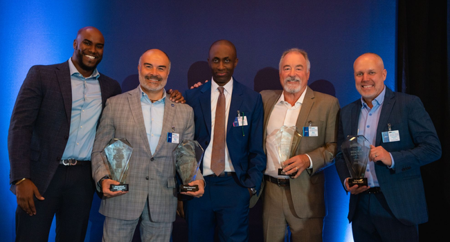 Group photo of five men in suits, smiling and holding four large glass awards won at this year's JetBlue business awards. Group photo of five men in suits, smiling and holding four large glass awards won at this year's JetBlue business awards.