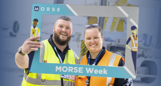 A man with short hair and a beard smiles alongside a girl with her hair tied back and glasses on her head. Both are wearing high visibility clothing and dressed for working on the airport ramp. They are holding a light blue and grey sign shaped like a picture frame  that features the words  A man with short hair and a beard smiles alongside a girl with her hair tied back and glasses on her head. Both are wearing high visibility clothing and dressed for working on the airport ramp. They are holding a light blue and grey sign shaped like a picture frame  that features the words