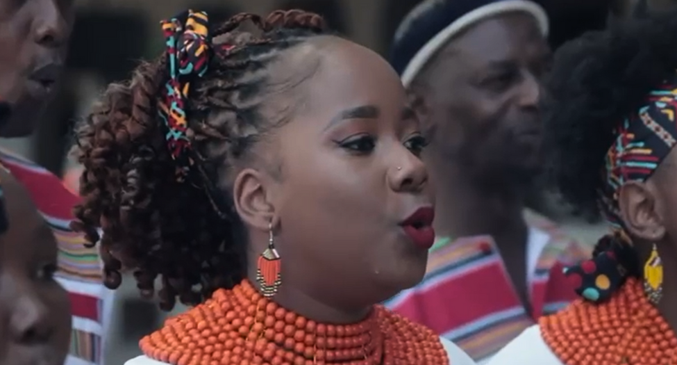 A lady singing wearing traditional dress and with braided hair featuring brightly coloured accessories. She is surrounded by people, also wearing traditional clothing, as they sing together in rows. the event celebrated the start of MORSE Week 2 at Menzies.