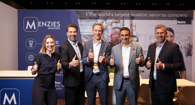 Five people (four men and one woman) standing in front of a Menzies Aviation event stand. All are wearing smart clothing and smiling while holding thumbs-up signs in celebration of this big moment. Five people (four men and one woman) standing in front of a Menzies Aviation event stand. All are wearing smart clothing and smiling while holding thumbs-up signs in celebration of this big moment.